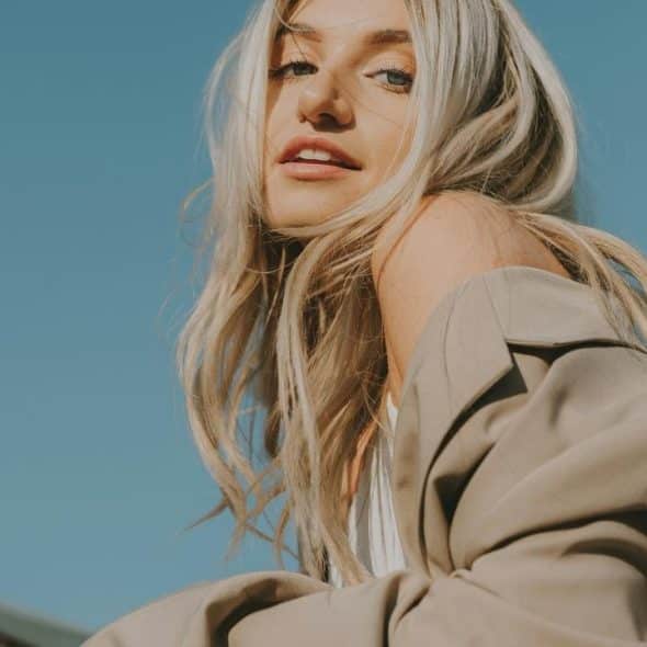 A woman with long blonde hair posing outdoors under a clear blue sky.