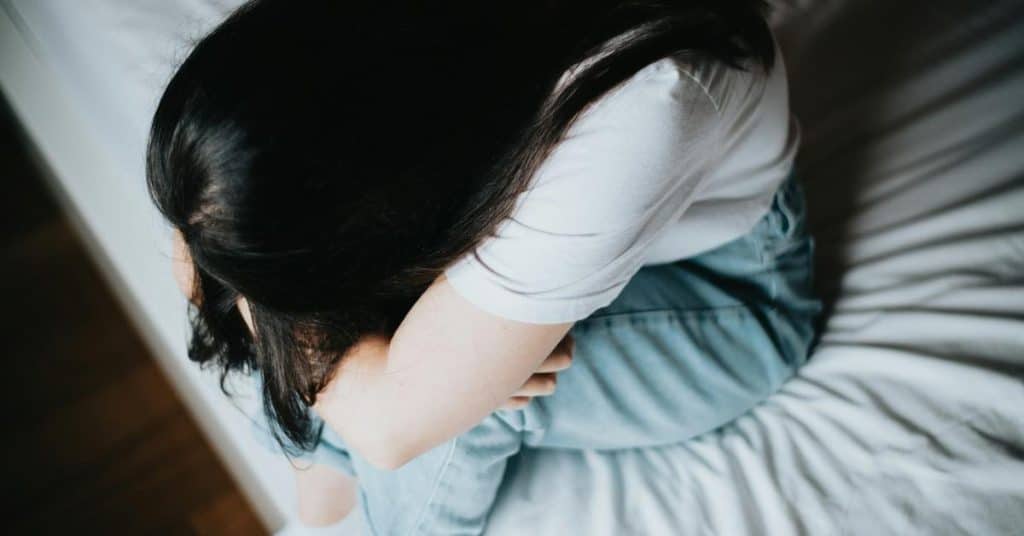 A person sitting curled up on a bed with their head down.