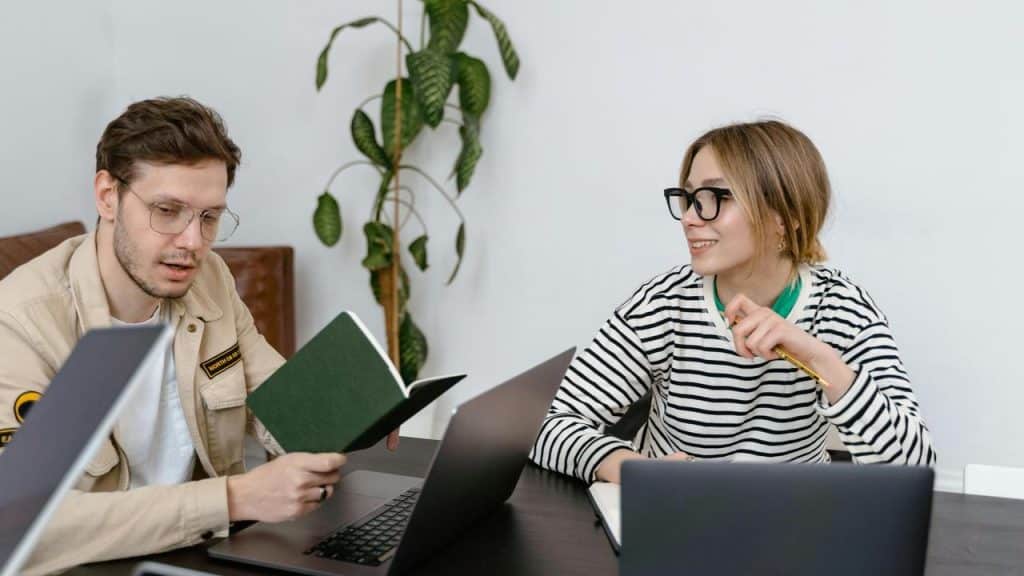 A man and woman working at a table with laptops and a notebook.