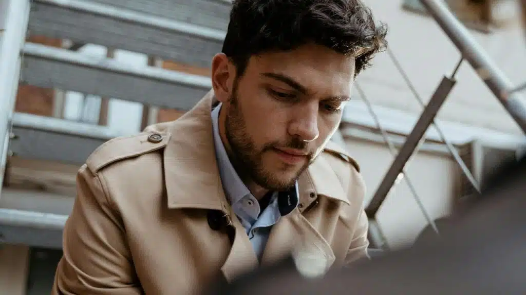 A man in a beige trench coat looking down while sitting on outdoor stairs.