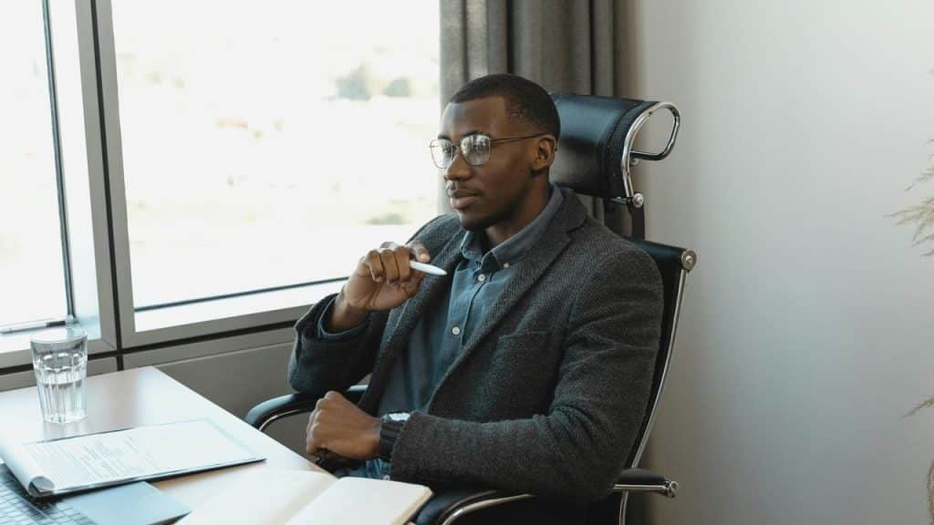 A man with glasses sitting thoughtfully at a desk by a window.
