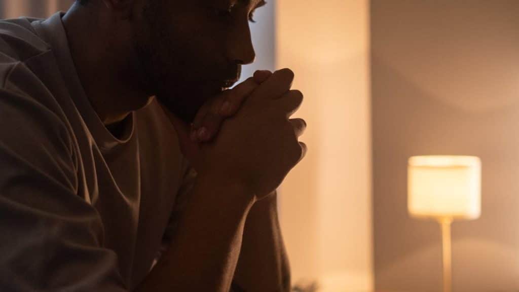 A man sitting with hands clasped in thought beside a lamp.