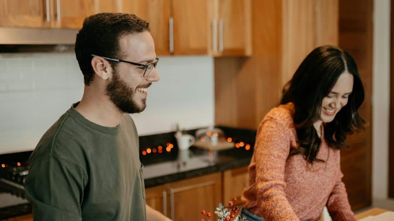 A couple smiling while preparing something together in a kitchen.