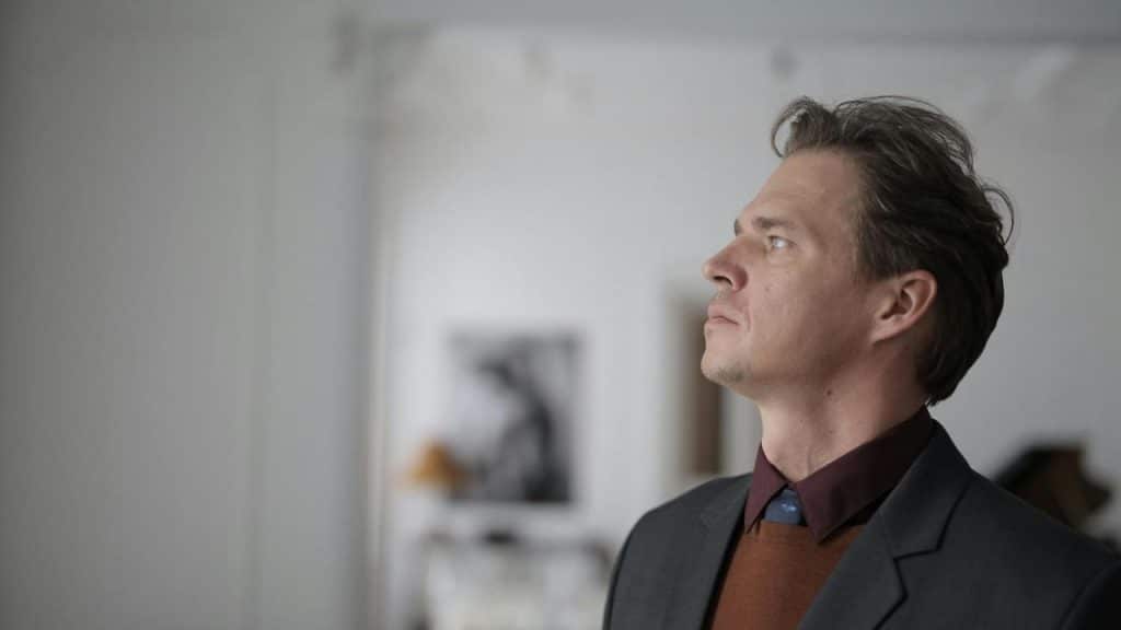 A man in a suit looking upward indoors with a blurred background.