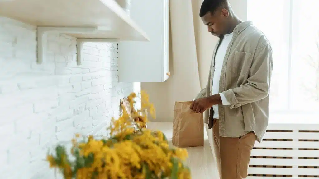 A man standing in a kitchen opening a paper bag on the counter.