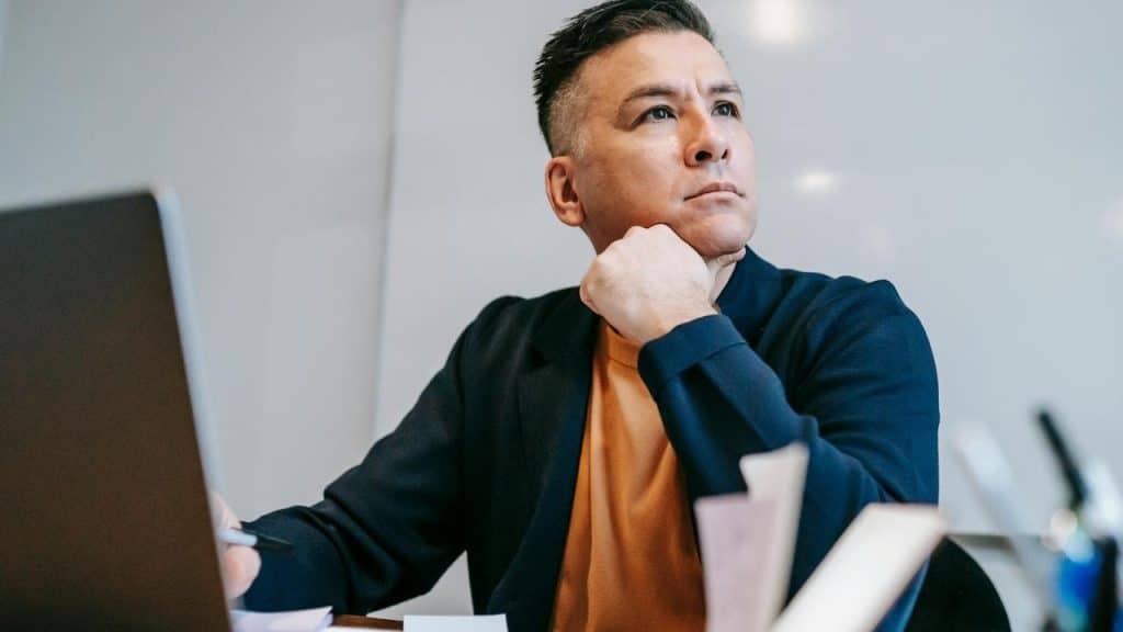 A man sitting at a desk with a laptop, looking upward thoughtfully.