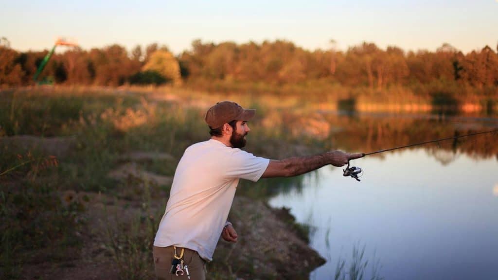 A man casts a fishing line by a lake at sunset.
