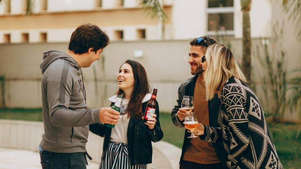 A group of friends stand outdoors talking and holding drinks.