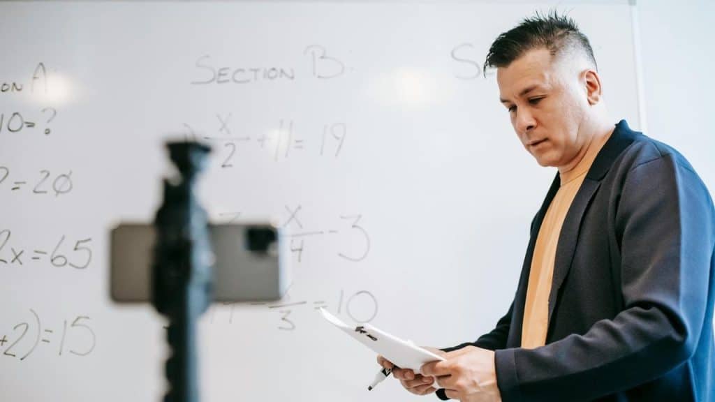 A teacher stands in front of a whiteboard holding papers.