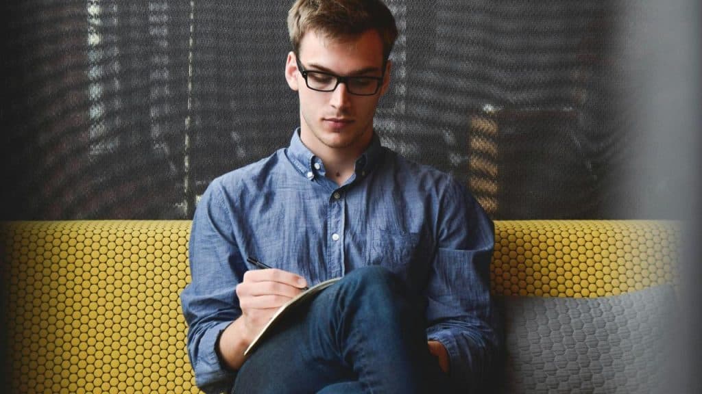 A man wearing glasses writes in a notebook while sitting on a couch.