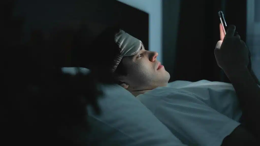 A man lying in bed at night looks at his phone while wearing a sleep mask.