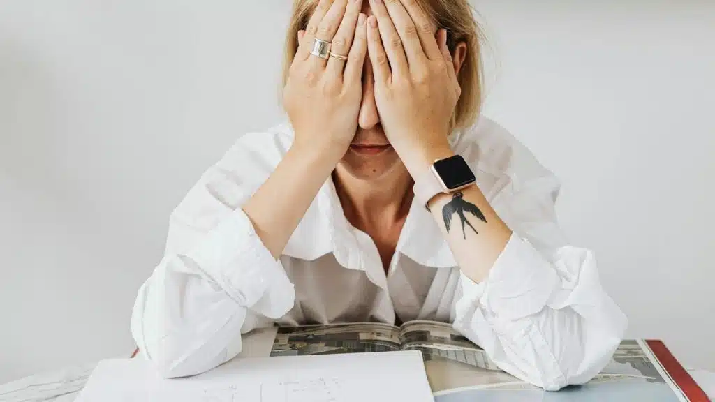 A woman covers her face with her hands while sitting at a desk with papers.