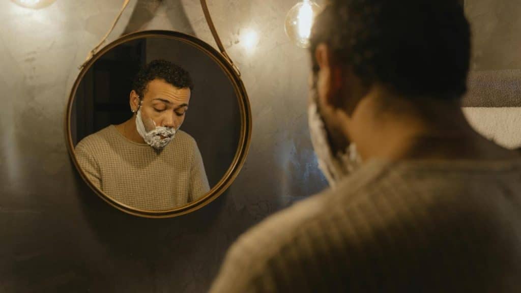 A man shaves his face while looking into a bathroom mirror.