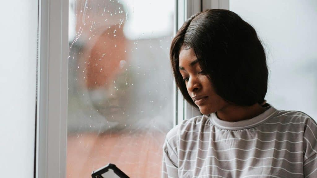 A woman looks at her phone while sitting by a rainy window.