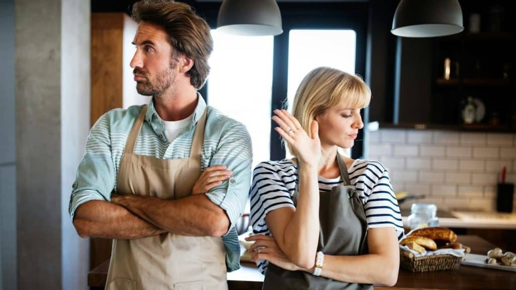 A man and woman wearing aprons stand back-to-back in a kitchen with arms crossed.