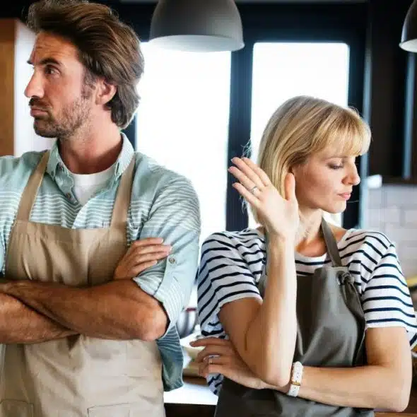 A man and woman wearing aprons stand back-to-back in a kitchen with arms crossed.