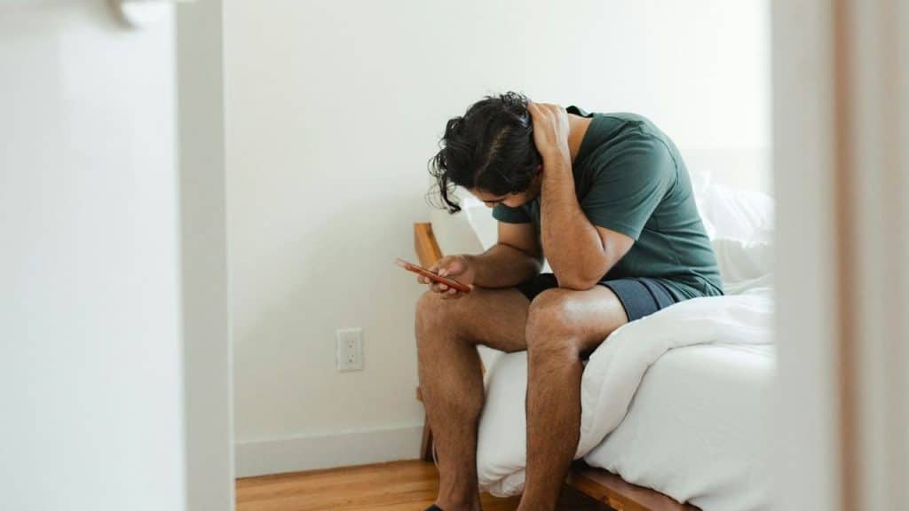 A man sits on a bed with one hand on his neck while looking at a smartphone.