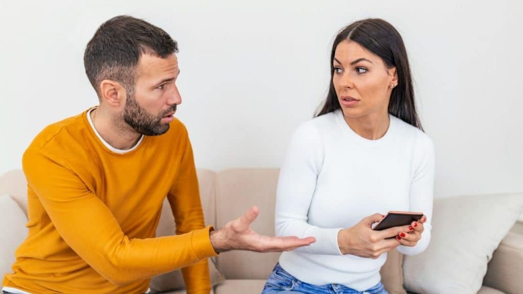 A man gestures toward a woman sitting on a sofa while she holds a smartphone.