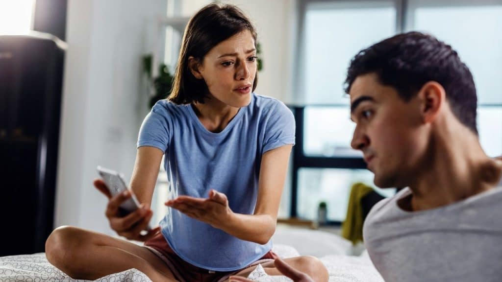 A woman holds a smartphone and gestures toward a man who is looking at her.