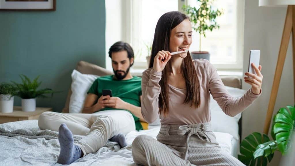 A woman brushes her teeth while taking a selfie near a man using a smartphone.