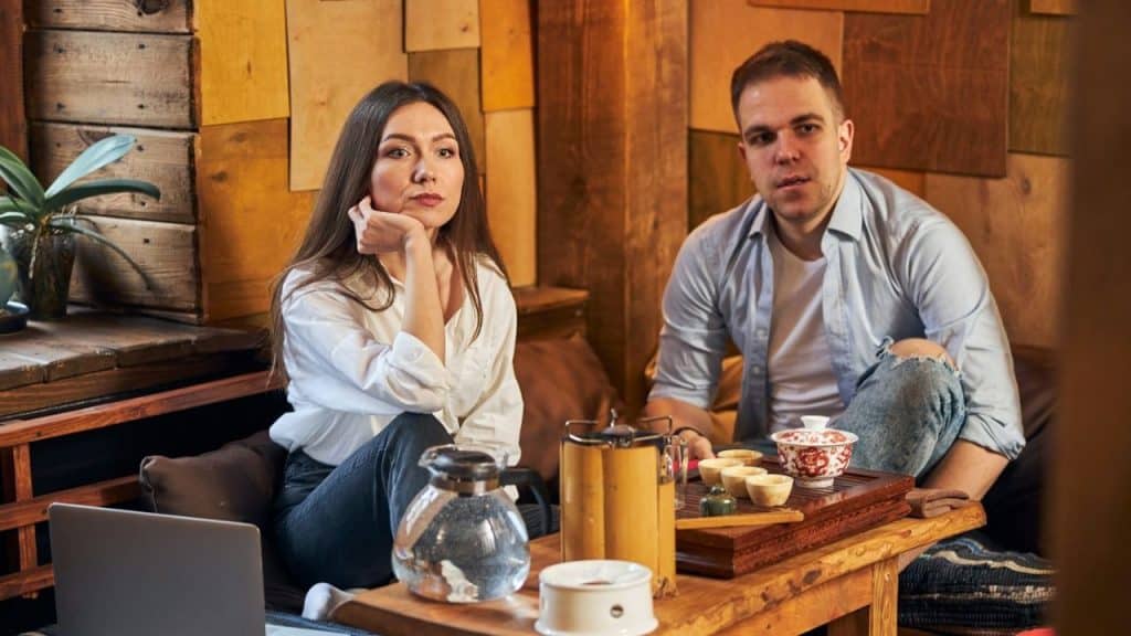 A woman and man sit behind a low wooden table featuring a tea set.
