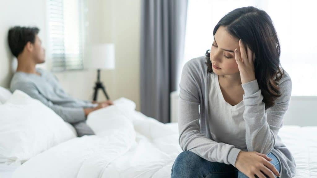 A woman sits on a bed touching her forehead while a man sits blurred behind.
