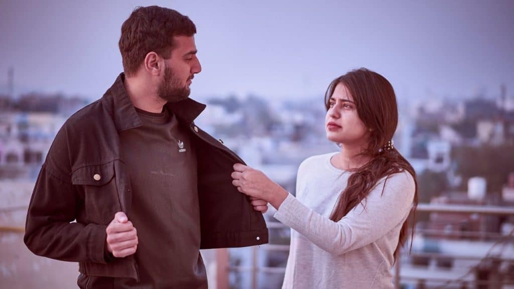 A woman stands outside holding a man’s jacket while they look at one another.