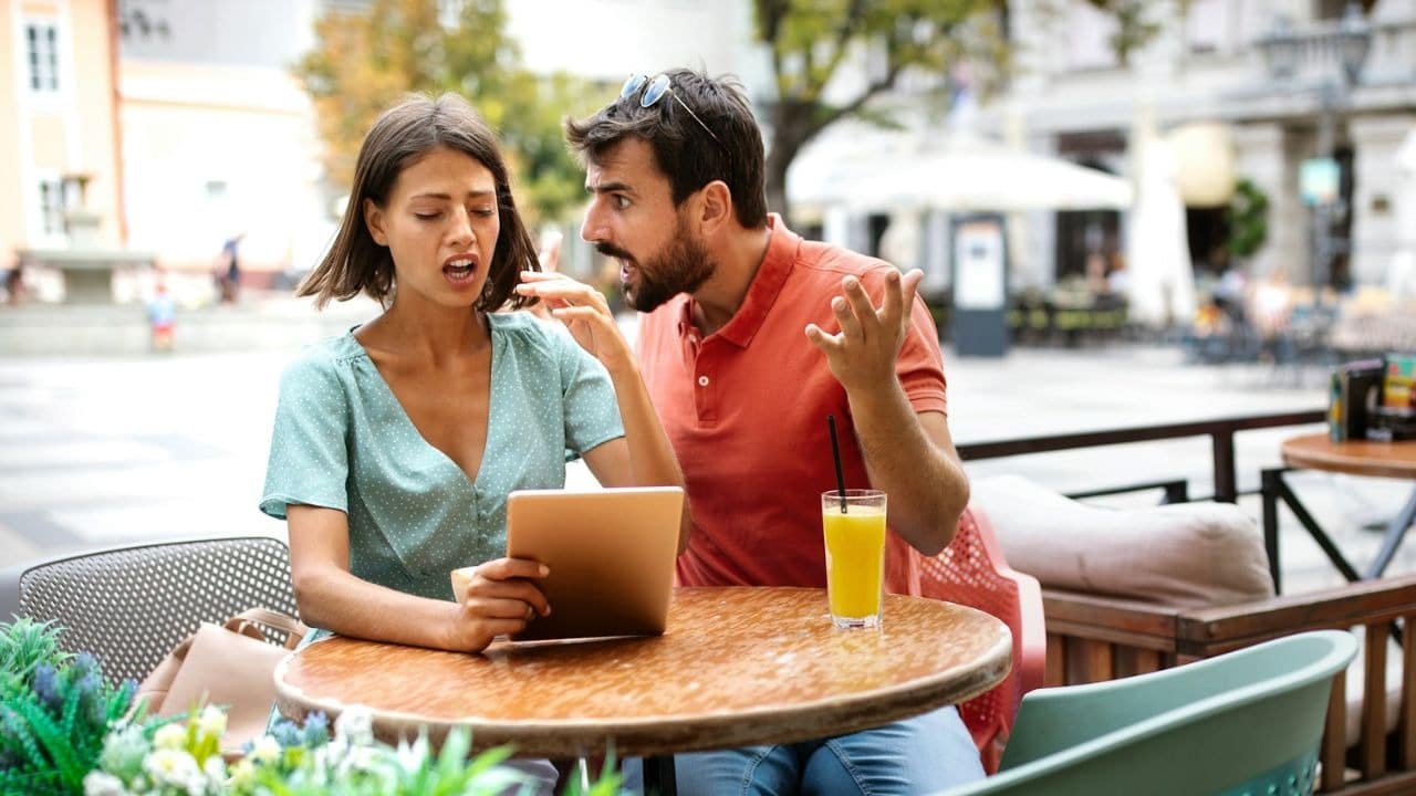 A man speaks emphatically with hand gestures toward a woman holding a tablet at a cafe.