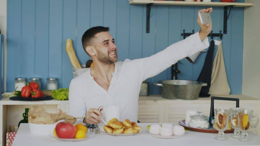 A smiling man sits at a kitchen table holding a smartphone up to take a selfie.
