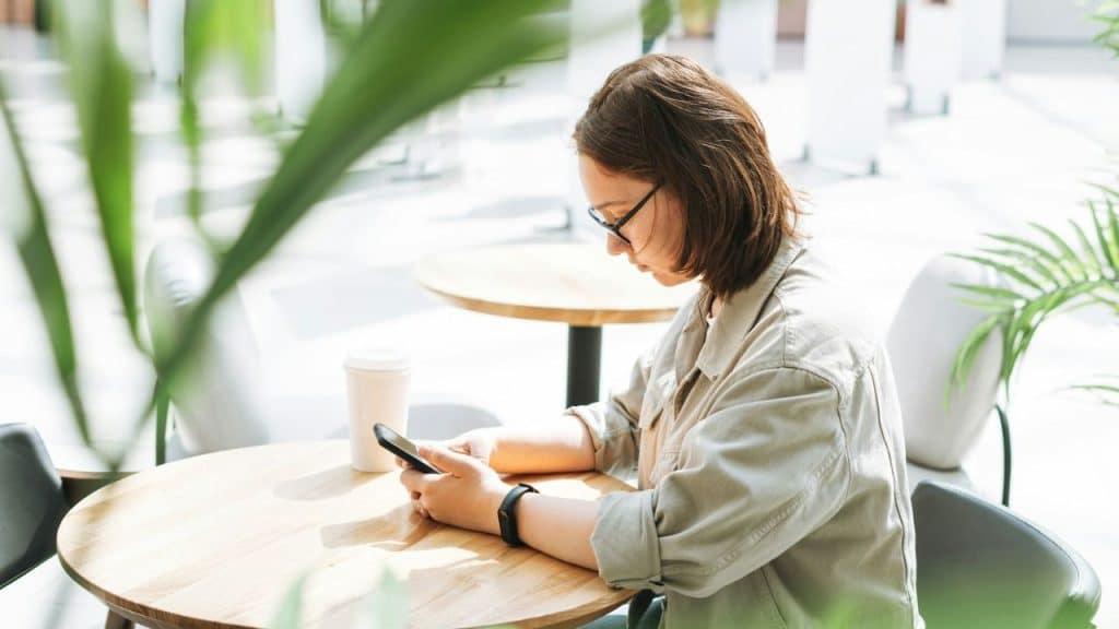 A person wearing glasses sits at an outdoor table looking down at a handheld smartphone.