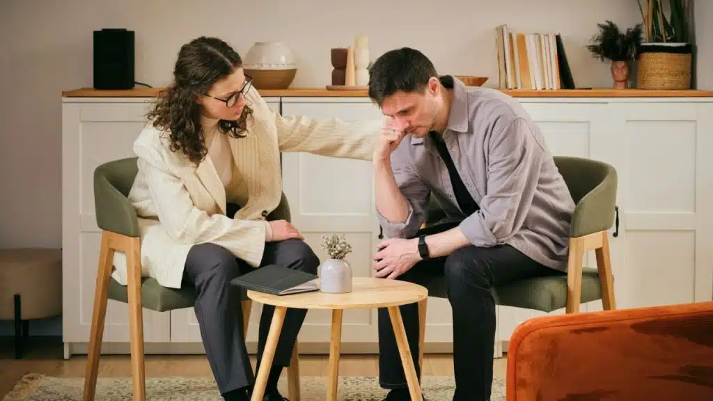 A woman in a blazer places her hand on the shoulder of a seated man.