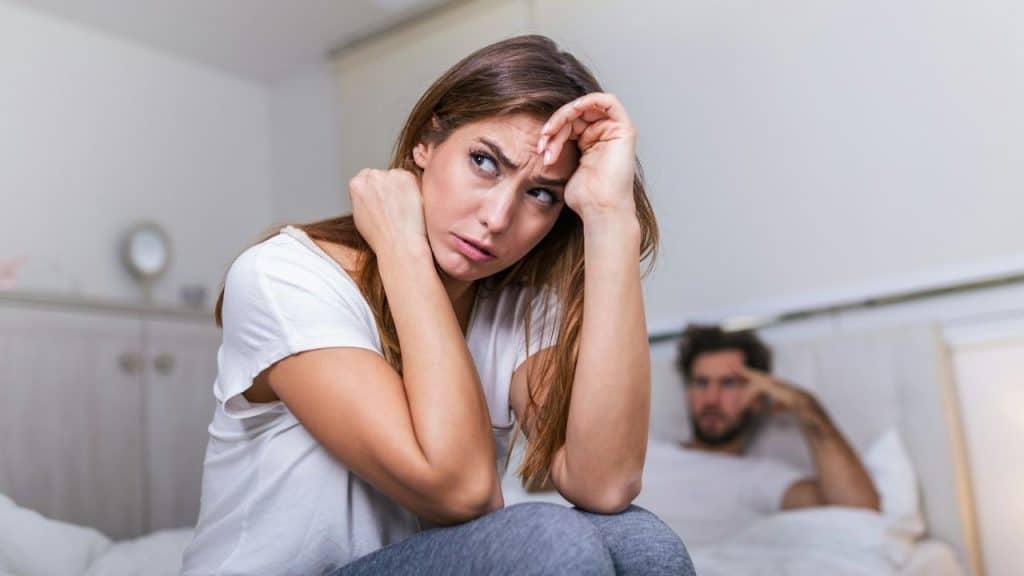 A woman with a distressed expression sits on a bed while a man watches behind.