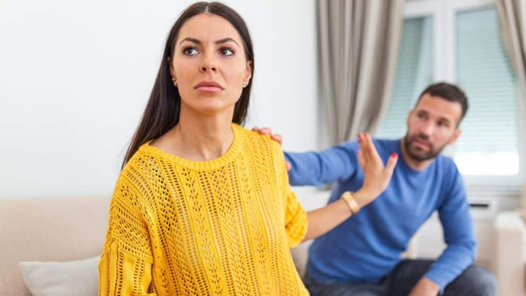 A woman in a yellow sweater looks away while a man reaches toward her shoulder.