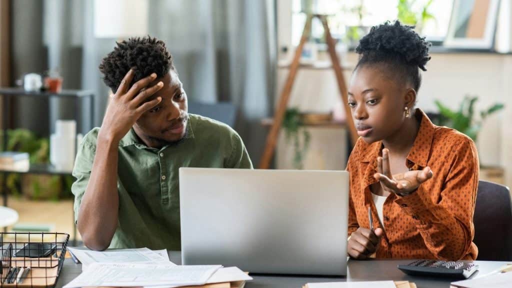 A man and woman sit at a desk looking at a laptop with various documents.