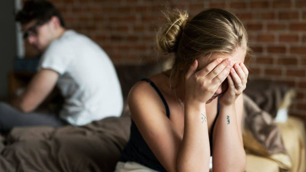 A woman sits on a bed with her face buried in her hands beside a man.
