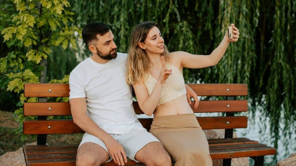 A woman holds up a smartphone to take a selfie with a man on a bench.