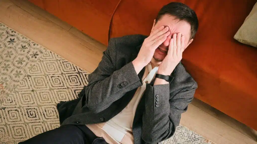 A man sits on the floor with his hands covering his face near a couch.