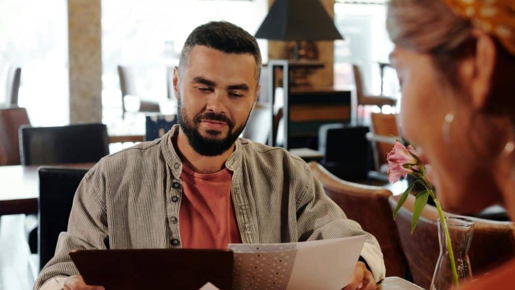 A man with a beard sits at a restaurant table, looking down at a paper menu.