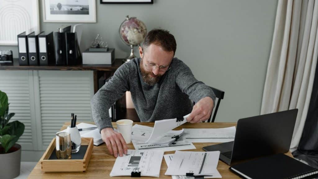 A man with glasses and a beard organizes various papers on a wooden office desk.