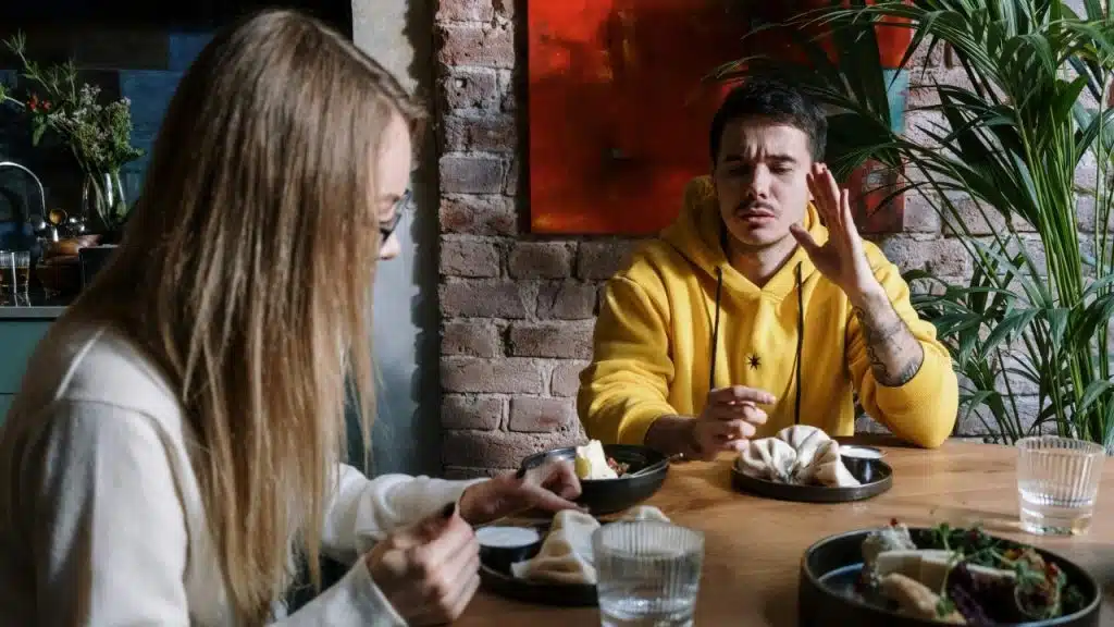 A man in a yellow hoodie sits at a table, gesturing while talking to someone.