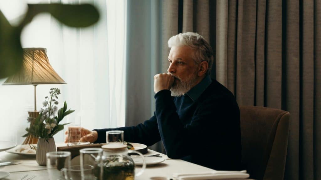 A man with white hair and a beard sits at a table, looking out a window.