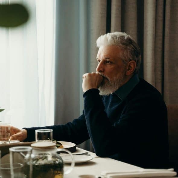 A man with white hair and a beard sits at a table, looking out a window.