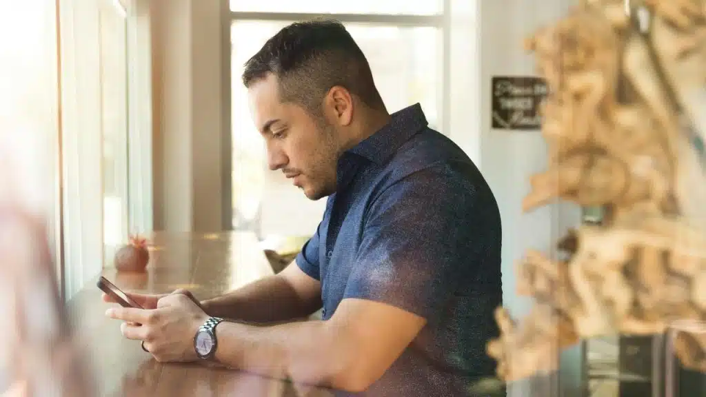 A man in a blue shirt leans on a wooden counter while looking at a smartphone.