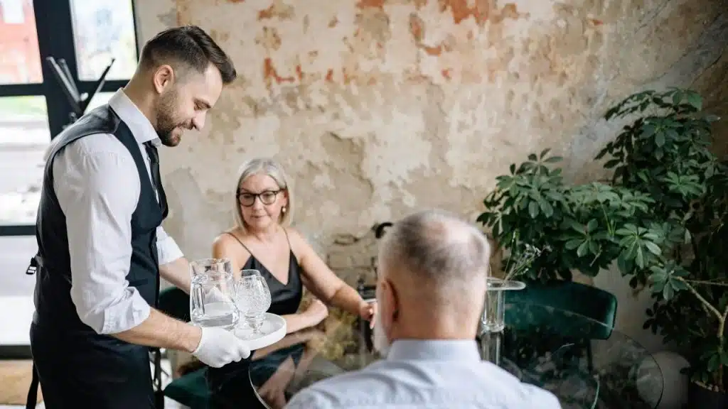 A server in a vest carries a tray with water to a couple at a table.