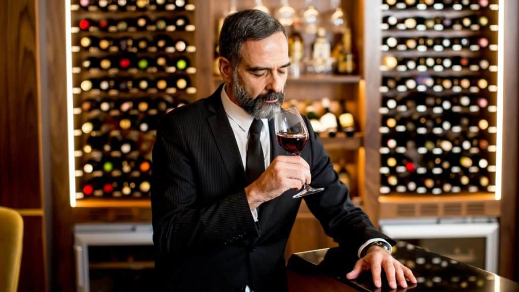 A man in a black suit holds a glass of red wine in a cellar.