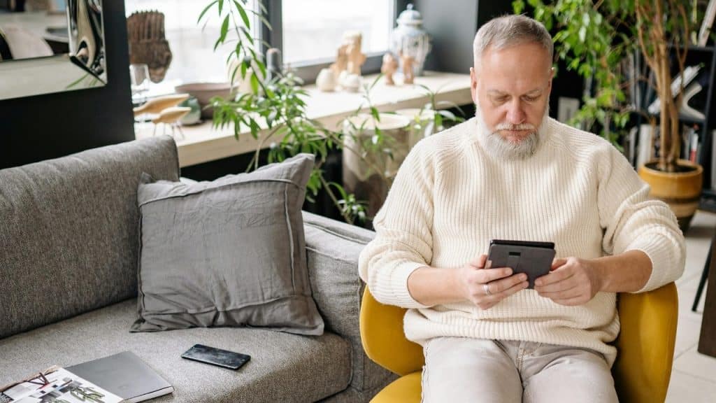 A man with a white beard sits in a yellow chair, holding a digital tablet.
