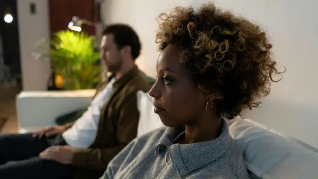 A woman with curly hair sits on a sofa next to a man, looking away.