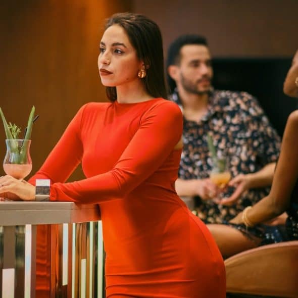 A woman in a red dress leans on a bar while two people sit behind her.