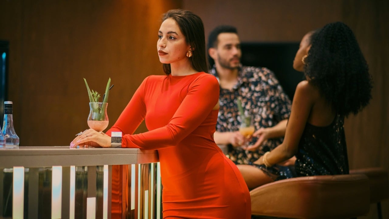 A woman in a red dress leans on a bar while two people sit behind her.