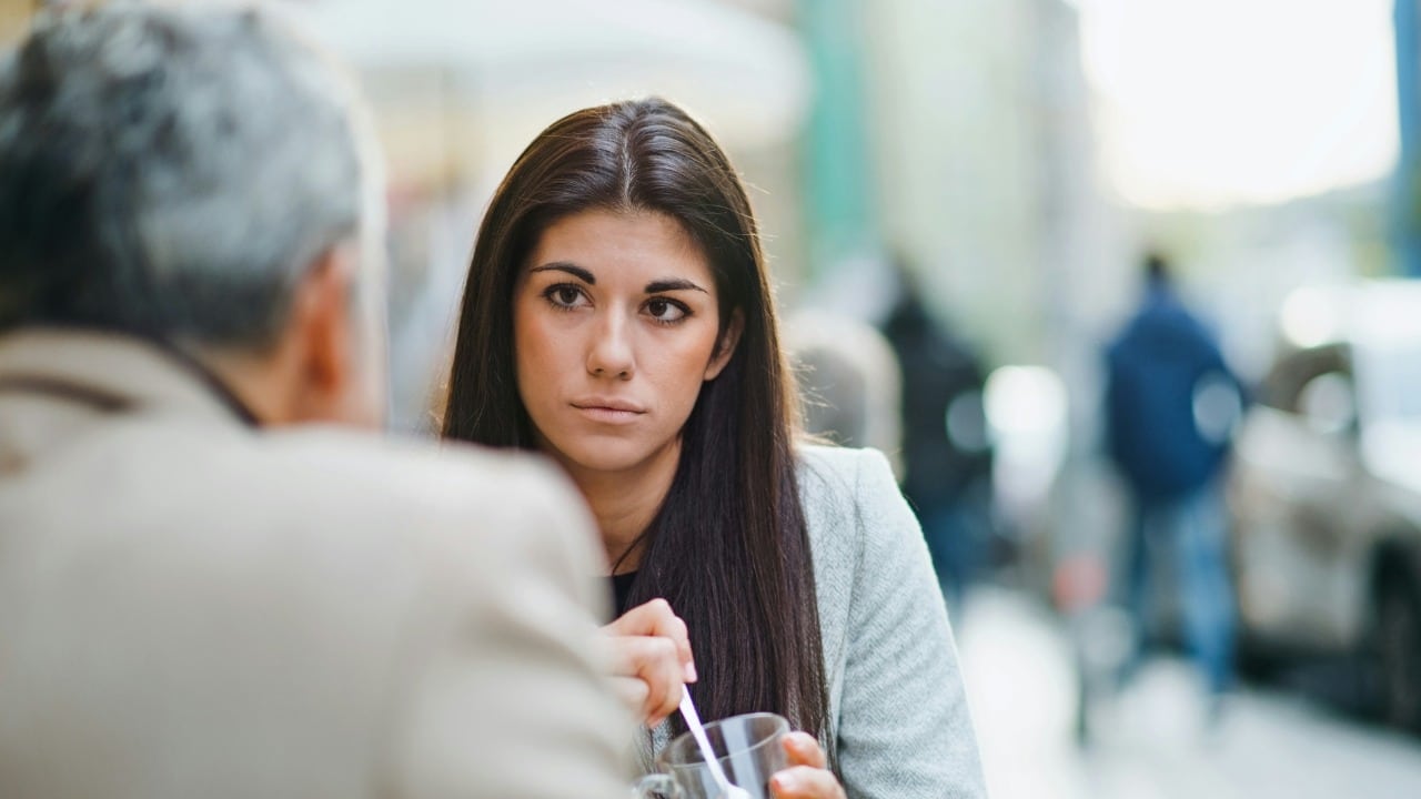 A woman with long dark hair looks toward a person whose back is to camera.